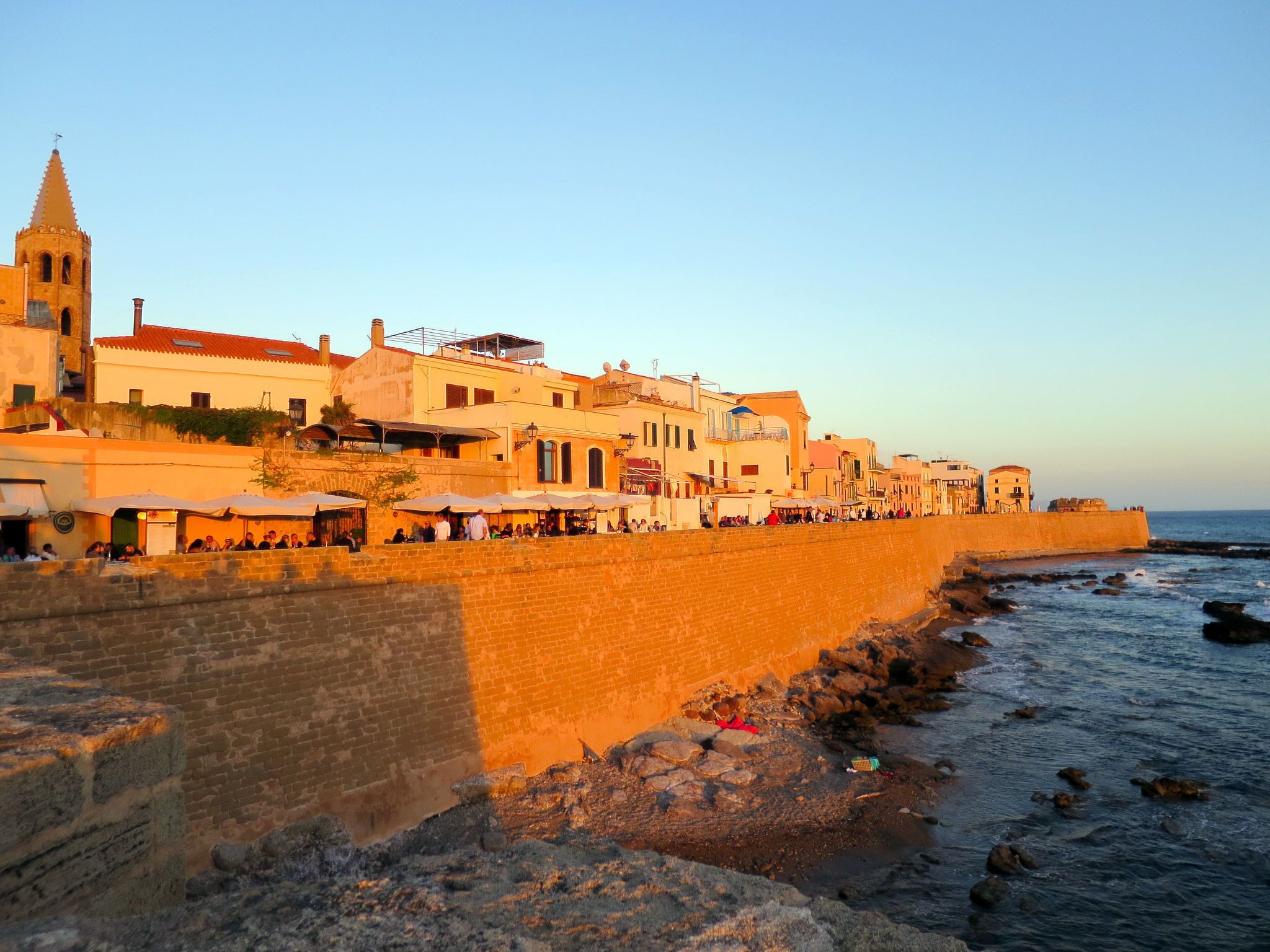 Une ville côtière qui se distingue par son imposant clocher d’église, un mur de pierre qui longe la plage et des bâtiments dotés de terrasses extérieures.