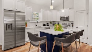 Cocina moderna con armarios blancos, electrodomésticos de acero inoxidable, una isla central con taburetes y luminarias colgantes.