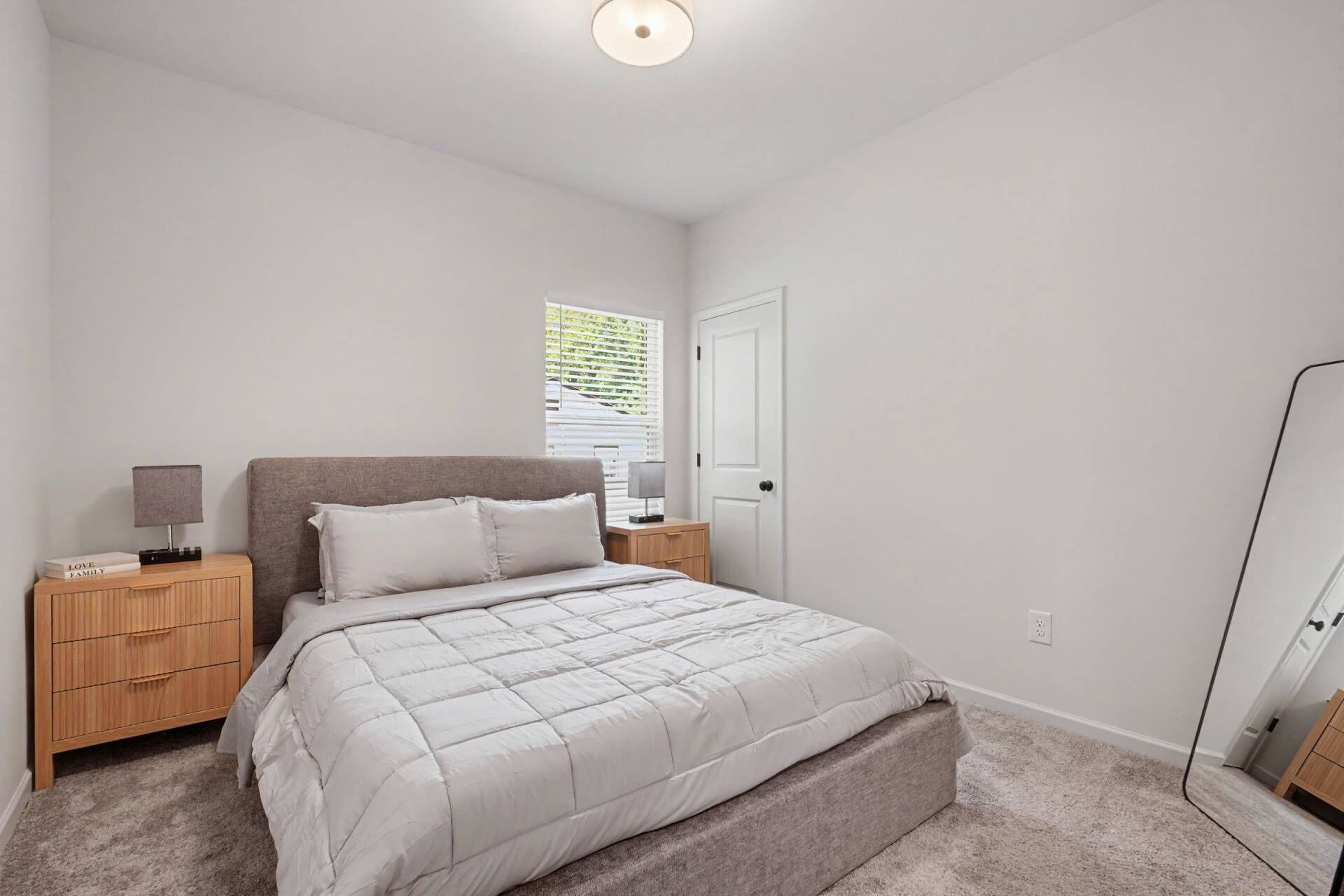 A bedroom with a bed, a nightstand, a lamp, a window with blinds, and a door.