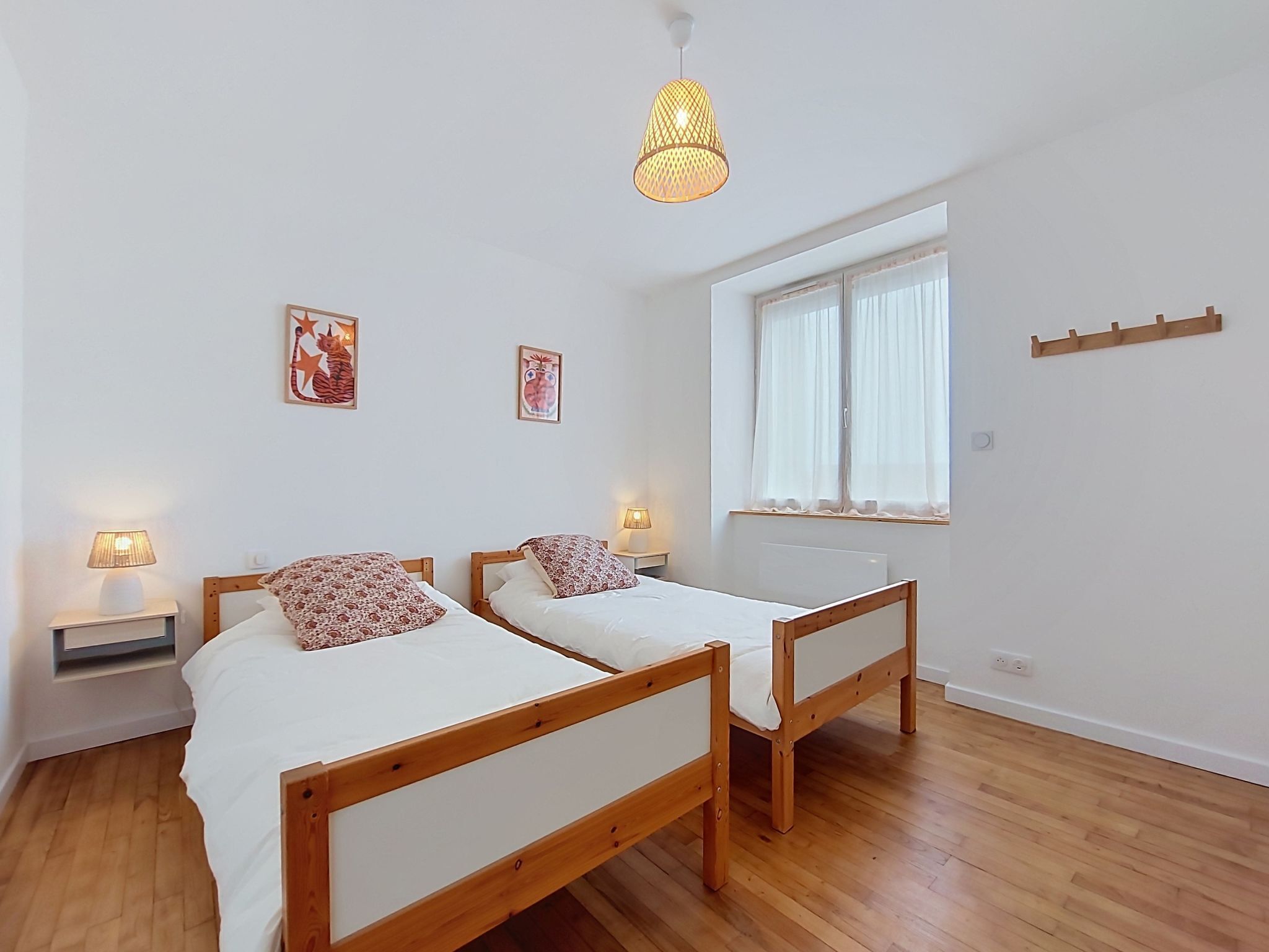 A room with two wooden beds, a window with curtains, a hanging light fixture, and wall-mounted artwork.