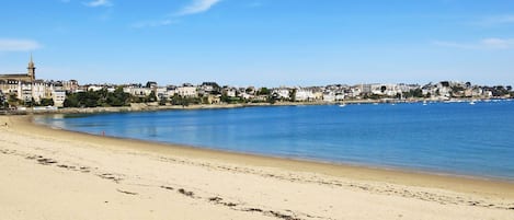 Une plage avec un ciel bleu dégagé, un rivage sablonneux et une ville dont les bâtiments se dessinent à l’arrière-plan.
