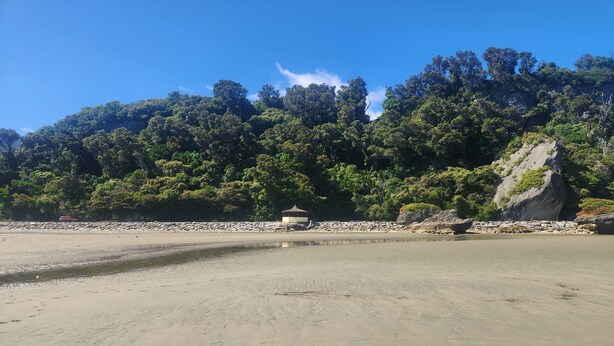 Una playa con un pabellón, cielos despejados y un bosque denso al fondo.