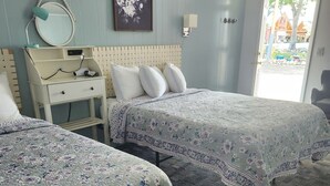 A hotel room with two beds, a bedside table with a lamp, a wall-mounted mirror, and a framed floral artwork.
