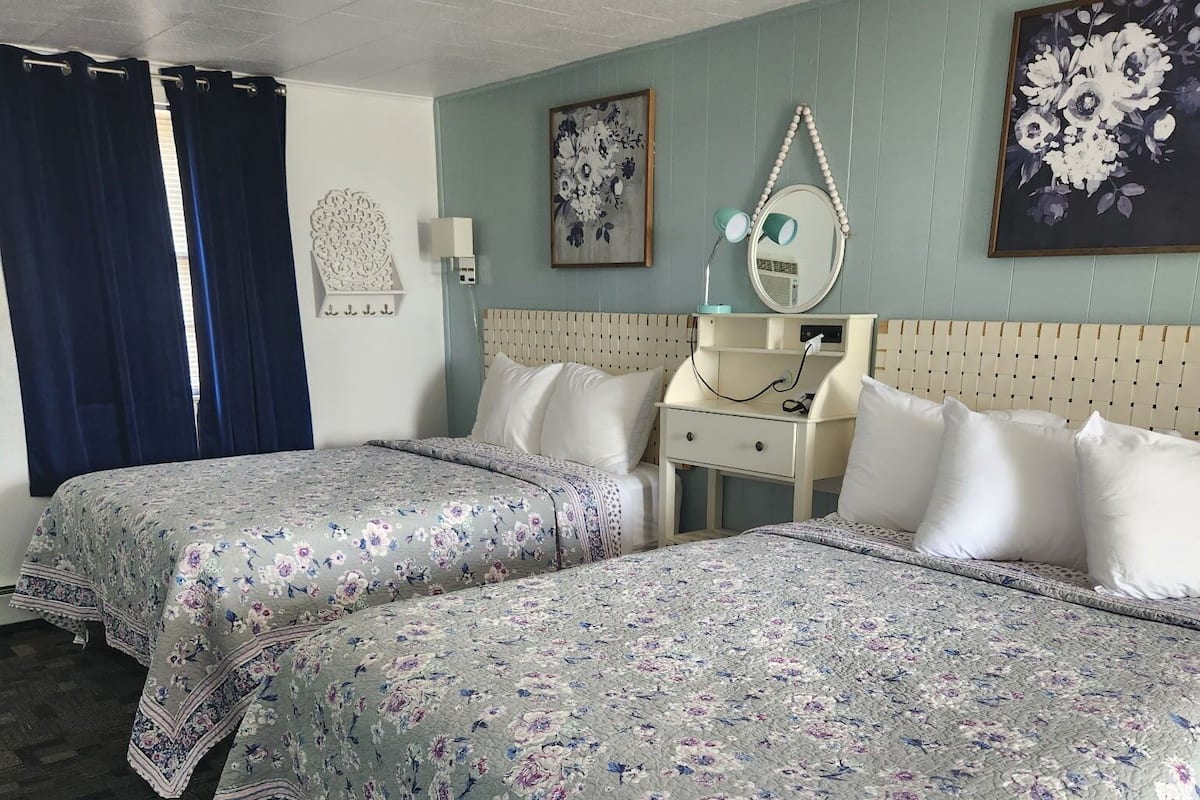 Een hotelkamer met twee bedden, een nachtkastje met een lamp, een spiegel en een bloemschilderij aan de muur.