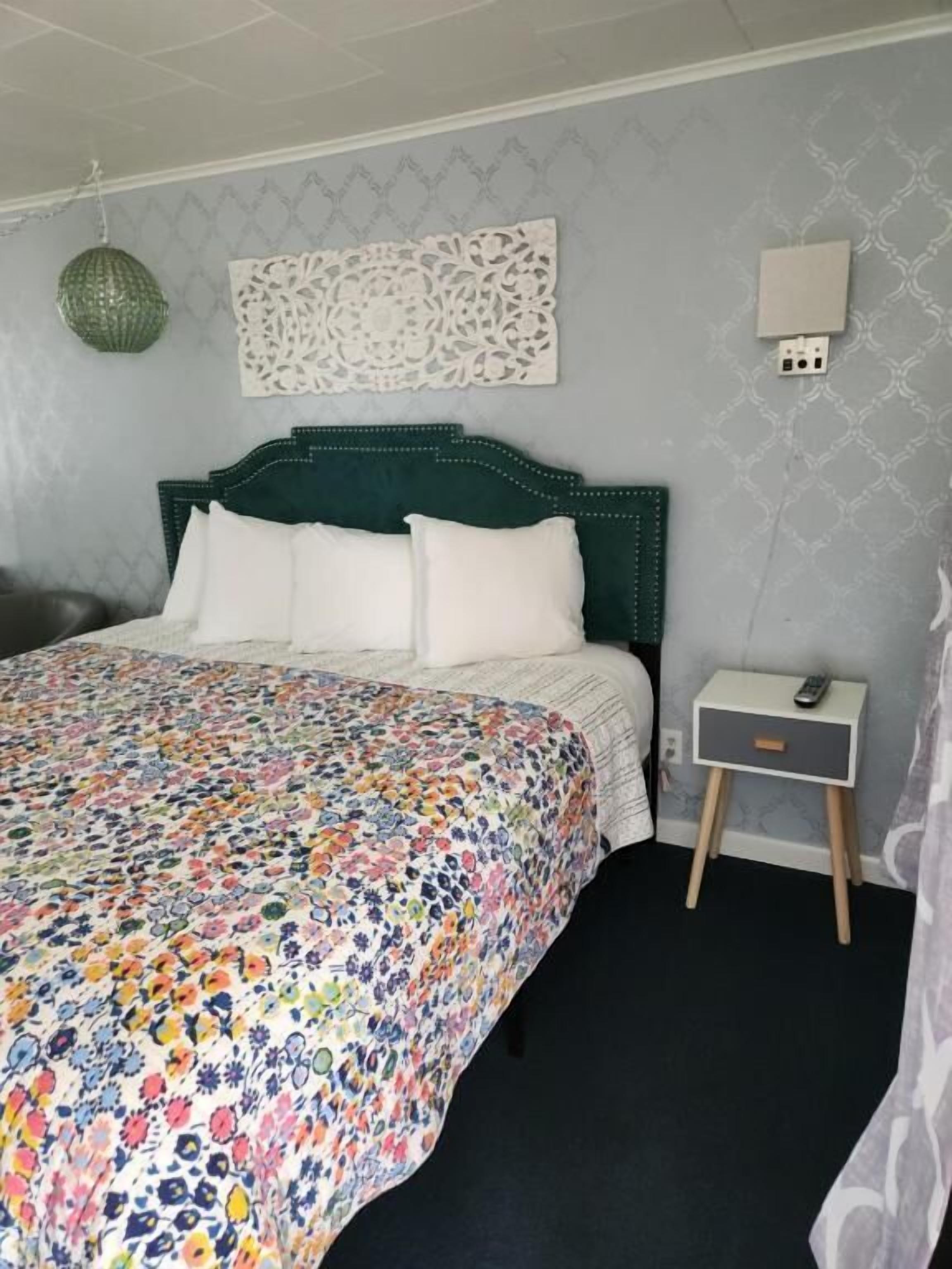 Une chambre à coucher avec une couvre-lit à motifs floraux, une tête de lit verte, une table de chevet avec une lampe et une prise de courant murale.