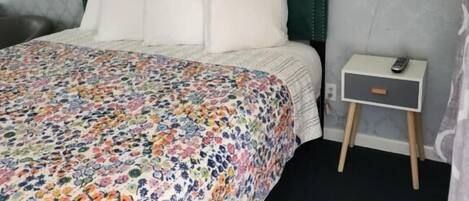 Une chambre à coucher avec une couvre-lit à motifs floraux, une tête de lit verte, une table de chevet avec une lampe et une prise de courant murale.