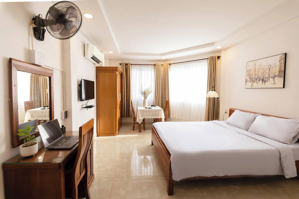 A hotel room with a bed, a desk with a laptop, a chair, a small table with a lamp, and a window with curtains.