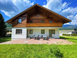 Ein zweistöckiges Holzhaus mit einem Balkon, einer Terrasse mit Tisch und Stühlen und Blick auf Berge im Hintergrund.