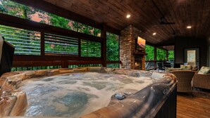 A hot tub with a stone fireplace, wooden ceiling, and outdoor seating area with a view of trees. - Luna Ridge · New! Cozy Escape: Hot Tub, Fire Pit, King Suite (Broken Bow)