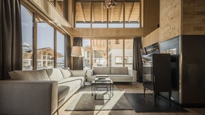 A spacious living room with a high ceiling, wooden beams, and large windows offering a view of the mountains. There is a sofa, a coffee table, and a flat-screen TV. - Luxury Chalet in Bichlbach (Bichlbach)