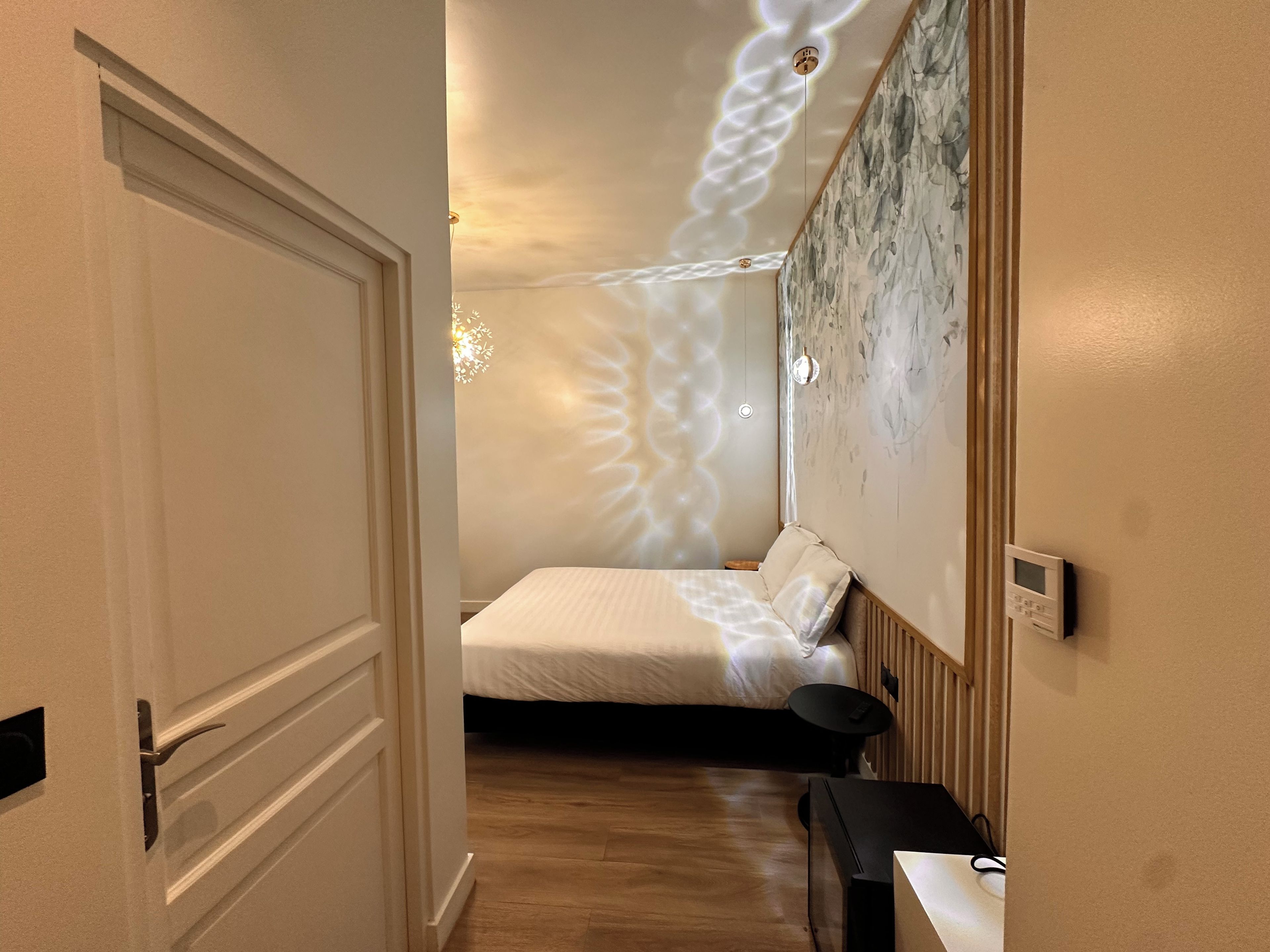 Une chambre à coucher comprenant un lit, une table de chevet, un thermostat fixé au mur et un plafond orné d’un luminaire original.