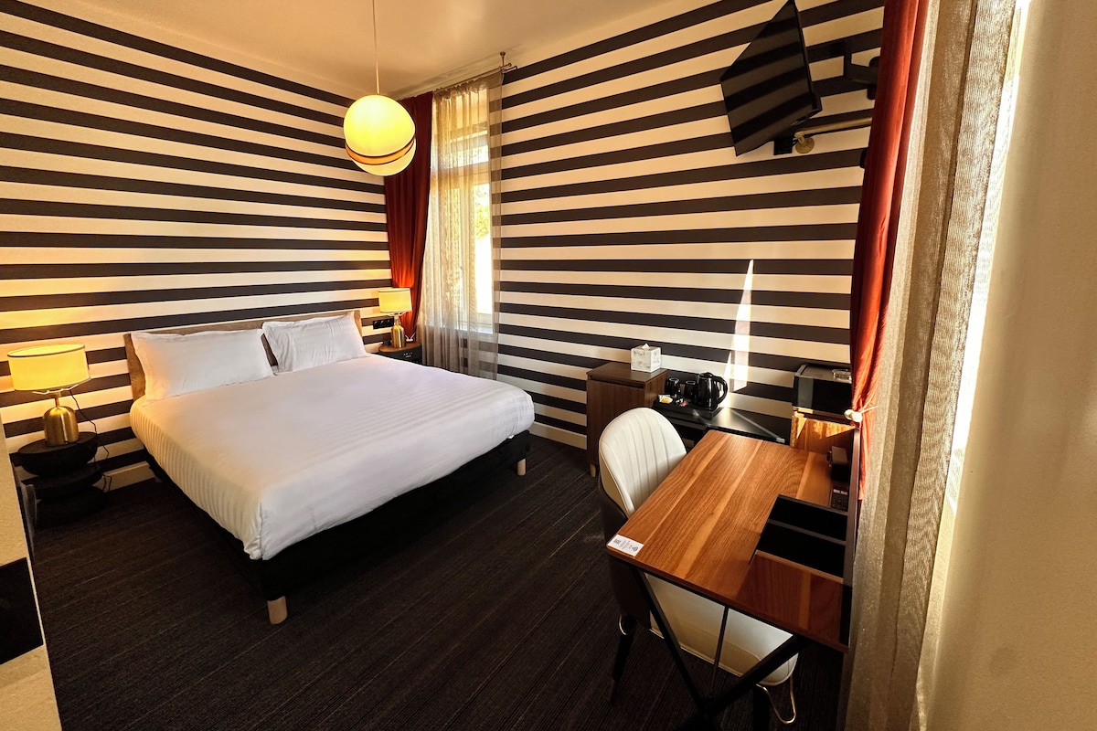 Ein Hotelzimmer mit Bett, Nachttisch, Lampe, Schreibtisch, Stuhl und einem wandmontierten Fernseher.