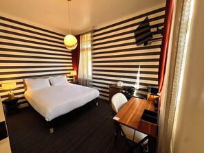 Habitación de hotel con cama, mesita de noche, lámpara, escritorio, silla y televisor de pared.
