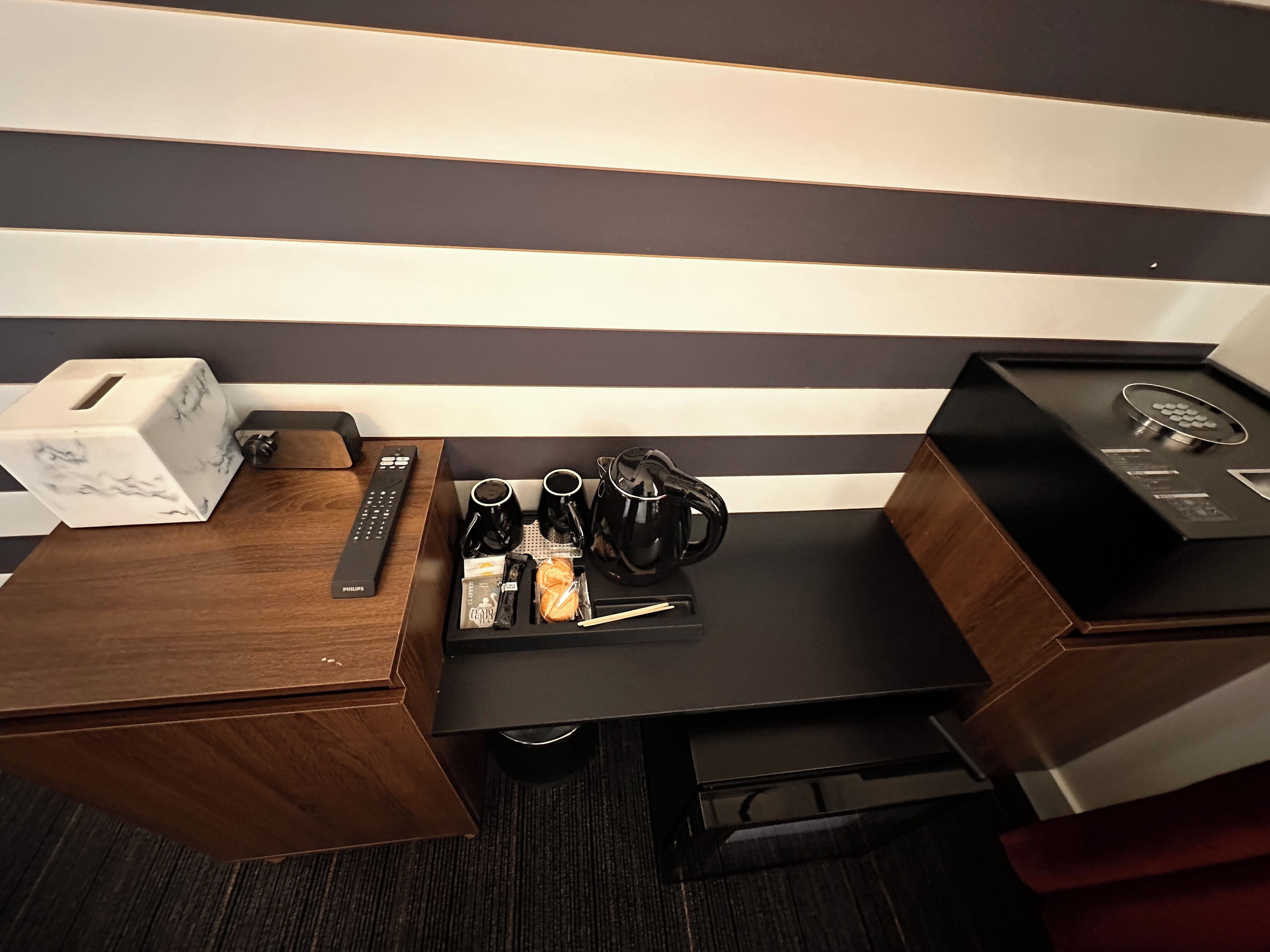 Une chambre d’hôtel avec un bureau en bois, une petite table basse noire, une boîte à motifs imitant le marbre, une télécommande, une machine à café, une bouilloire, une tasse et une petite assiette avec des en-cas.