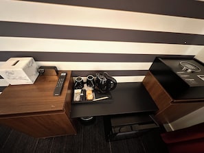 Une chambre d’hôtel avec un bureau en bois, une petite table basse noire, une boîte à motifs imitant le marbre, une télécommande, une machine à café, une bouilloire, une tasse et une petite assiette avec des en-cas.