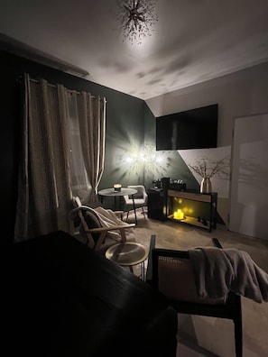 Ein schwach beleuchteter Raum mit einer Couch, einem kleinen runden Tisch, einem an der Wand befestigten Fernseher und einer dekorativen Leuchte.