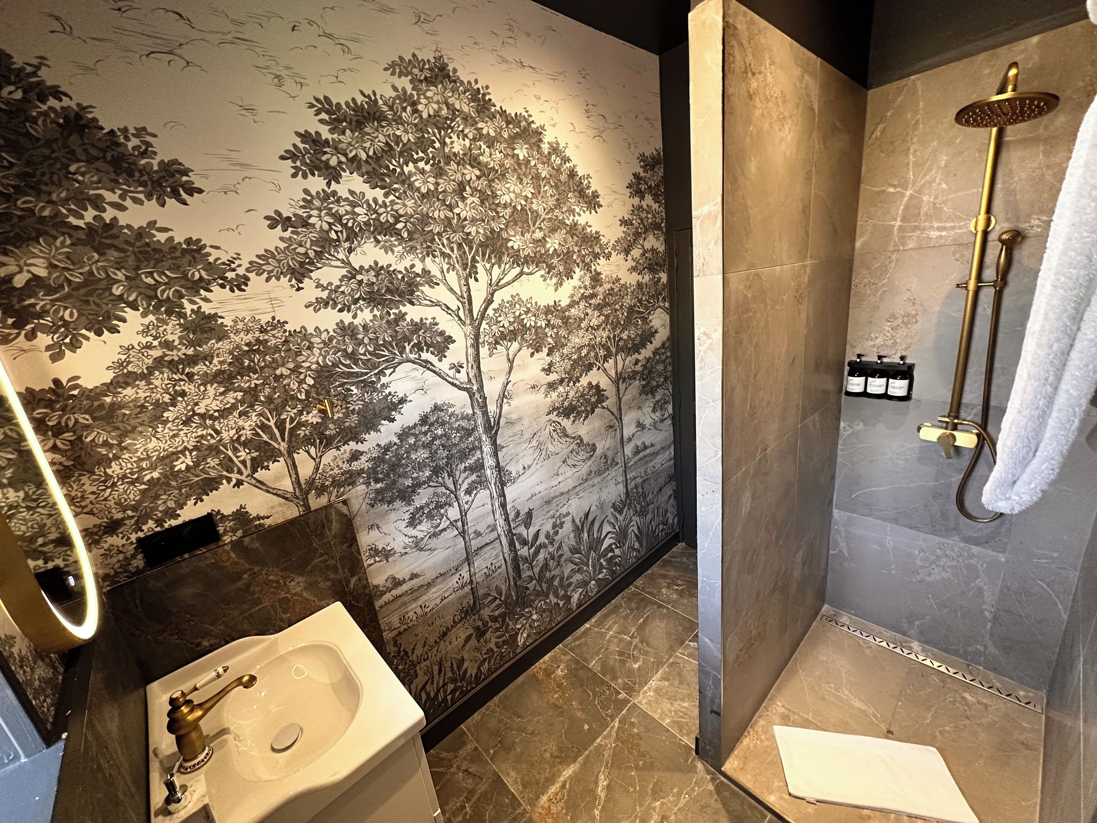 Een badkamer met een wastafel, een douche met een gouden douchekop en een muur met een muurschildering van een boom.