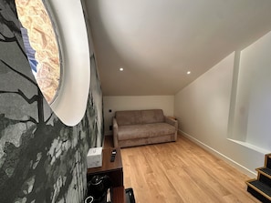 Un salon moderne avec un plancher en bois, un canapé beige et un mur en pierre agrémenté d’une fenêtre circulaire.
