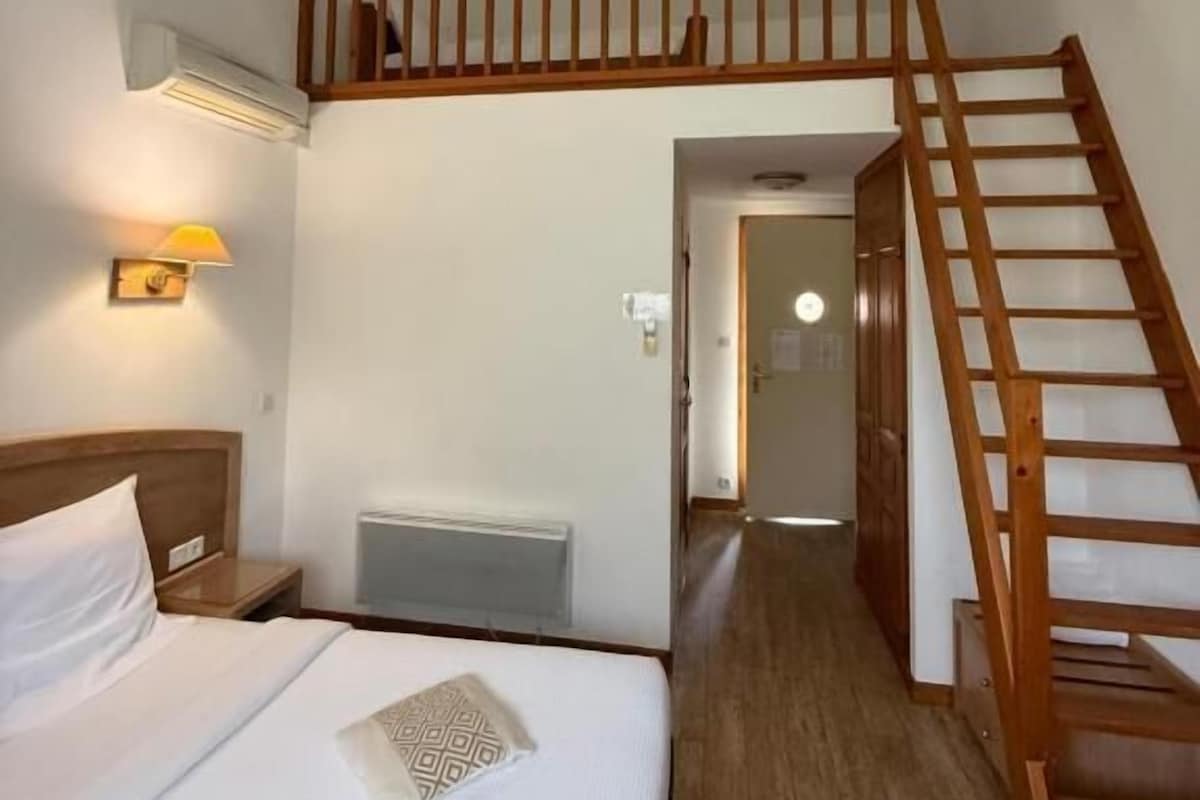 A bedroom with a bed, a nightstand, a wall-mounted heater, a wooden staircase, and a hallway leading to another room.