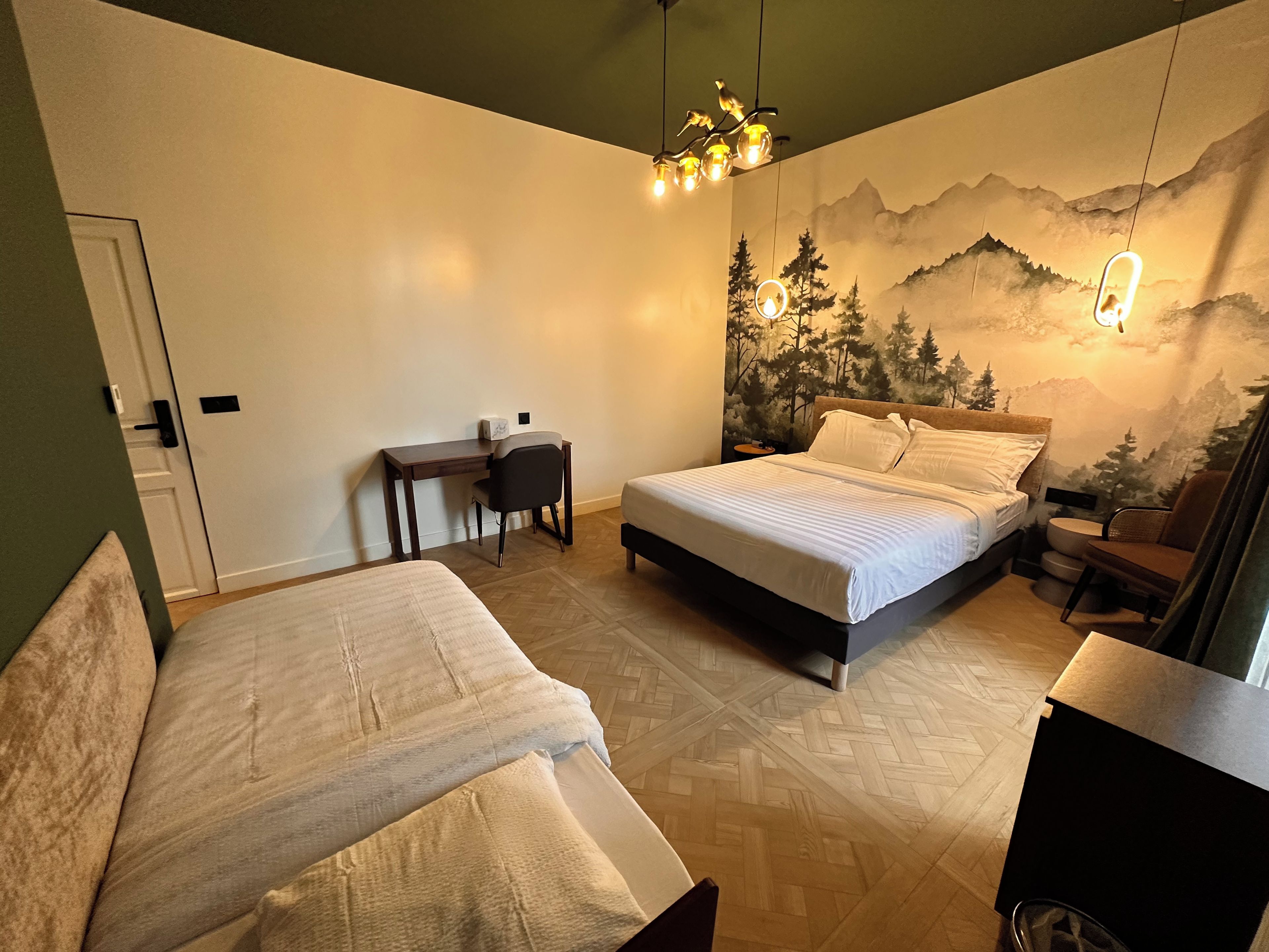 Een slaapkamer met een bed, een bureau en een stoel. Op de muur bevindt zich een muurschildering van een bos en bergen.