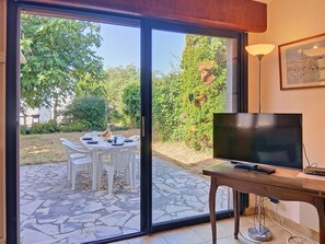 A room with a TV on a wooden table, a sliding glass door leading to an outdoor dining area with white plastic chairs and a table set for two, and a framed picture on the wall. - Arceau 3 by Interhome (Ile d'Oléron)