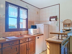 A kitchen with wooden cabinets, a white refrigerator, a microwave, and a window with patterned curtains. - Arceau 3 by Interhome (Ile d'Oléron)