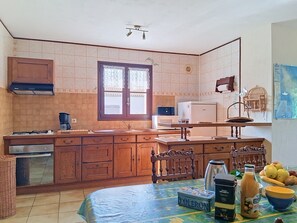A kitchen with wooden cabinets, a gas stove, a refrigerator, and a dining area with a table set for two. - Arceau 3 by Interhome (Ile d'Oléron)