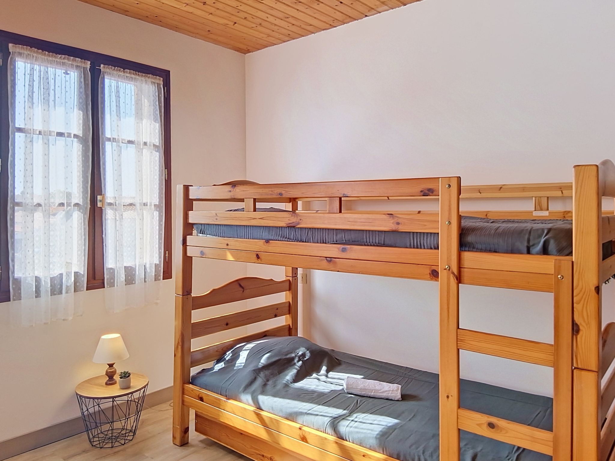 Una cama litera con estructura de madera, una mesita de noche pequeña con una lámpara y una ventana con cortinas traslúcidas.