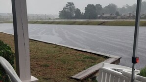 A covered outdoor area with white plastic chairs, a concrete floor, and a view of a wet road and distant trees.