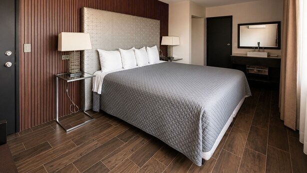 A hotel room with a large bed, bedside lamps, a nightstand, a dresser with a mirror, and a door. - Maviri Hotel (Tijuana)