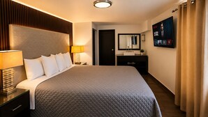 A hotel room with a large bed, a bedside table with a lamp, a dresser, a mirror, a TV, and a door. - Maviri Hotel (Tijuana)