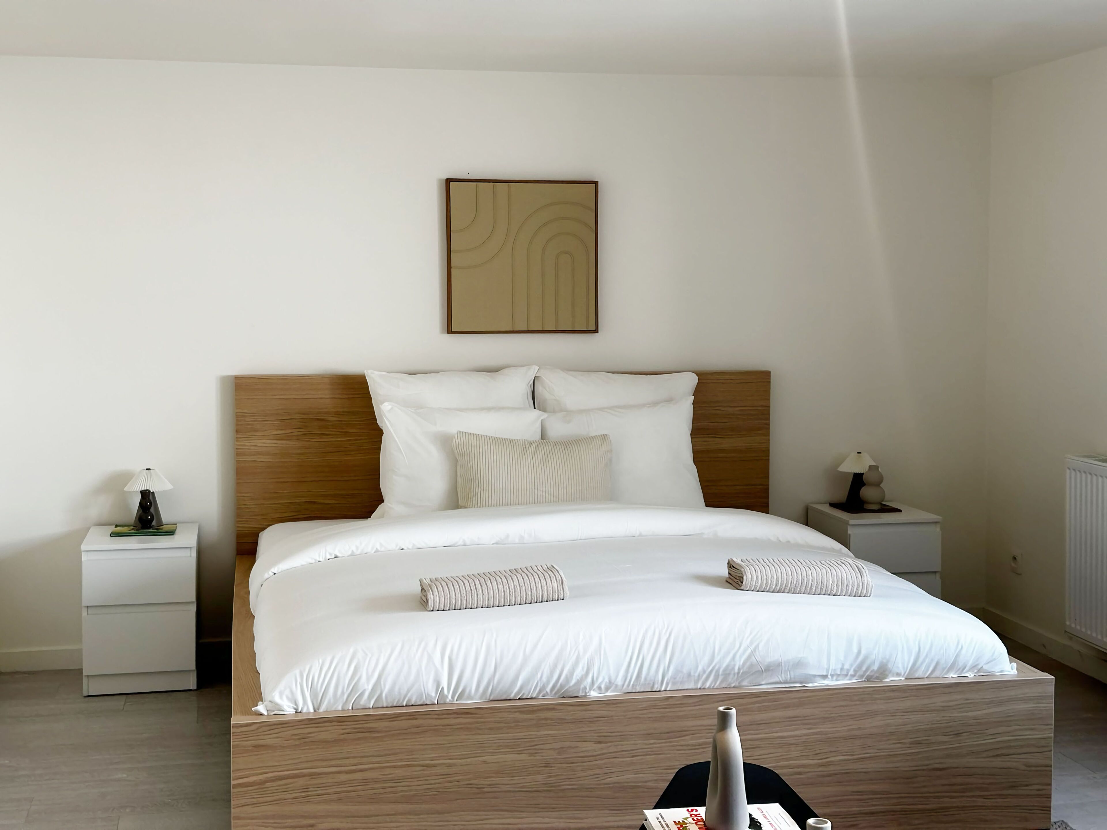 Una cama bien tendida con sábanas blancas, un cabecero de madera y dos mesitas de noche con lámparas.
