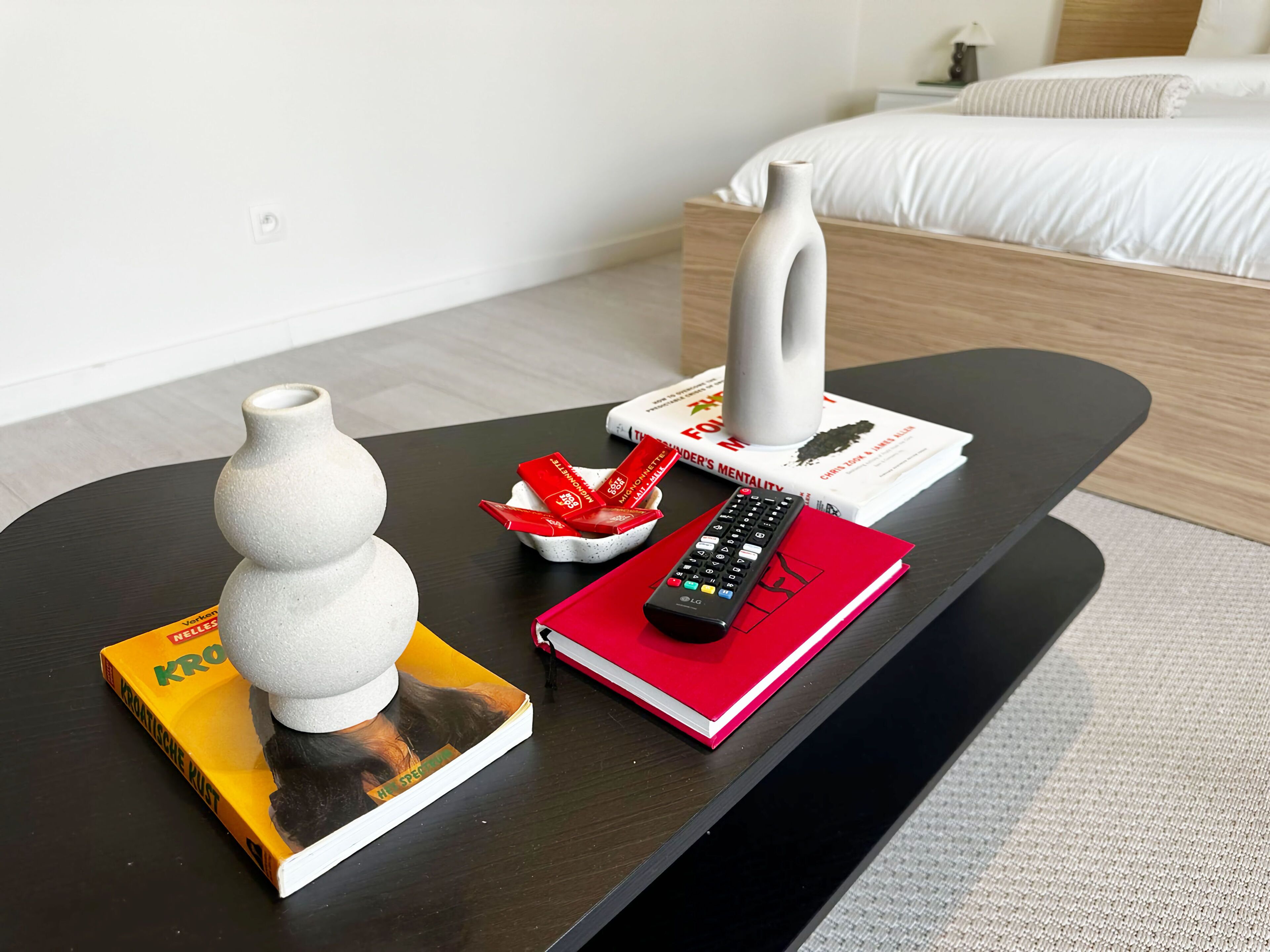 Habitació d'hotel amb llit, una taula de nit, un comandament a distància, un llibre, un ram i un bol d'aperitius.