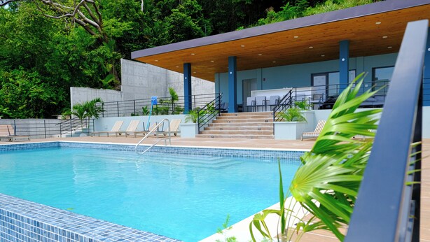 A swimming pool with a tiled edge, a wooden deck, and a covered seating area with chairs and a staircase. - Newly Built Apartment in the Heart of Ocho Rios (Ocho Rios)