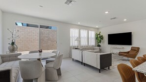 Uma sala de estar moderna com um conjunto de sofás, uma mesa de jantar com cadeiras, uma televisão na parede e uma parede de tijolos.