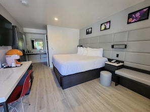 Ein modernes Hotelzimmer mit einem großen Bett, einem Schreibtisch mit Stuhl, einem kleinen Tisch, einer Mülleimer und einem an der Wand befestigten Fernseher.
