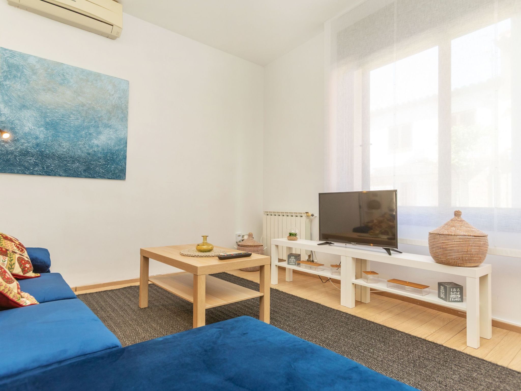 Una sala de estar moderna con un sofá azul, una mesa de centro de madera, un televisor sobre un mueble blanco y un ventanal con cortinas translúcidas.