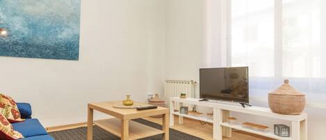 A modern living room with a blue sofa, a wooden coffee table, a TV on a white stand, and a large window with sheer curtains.