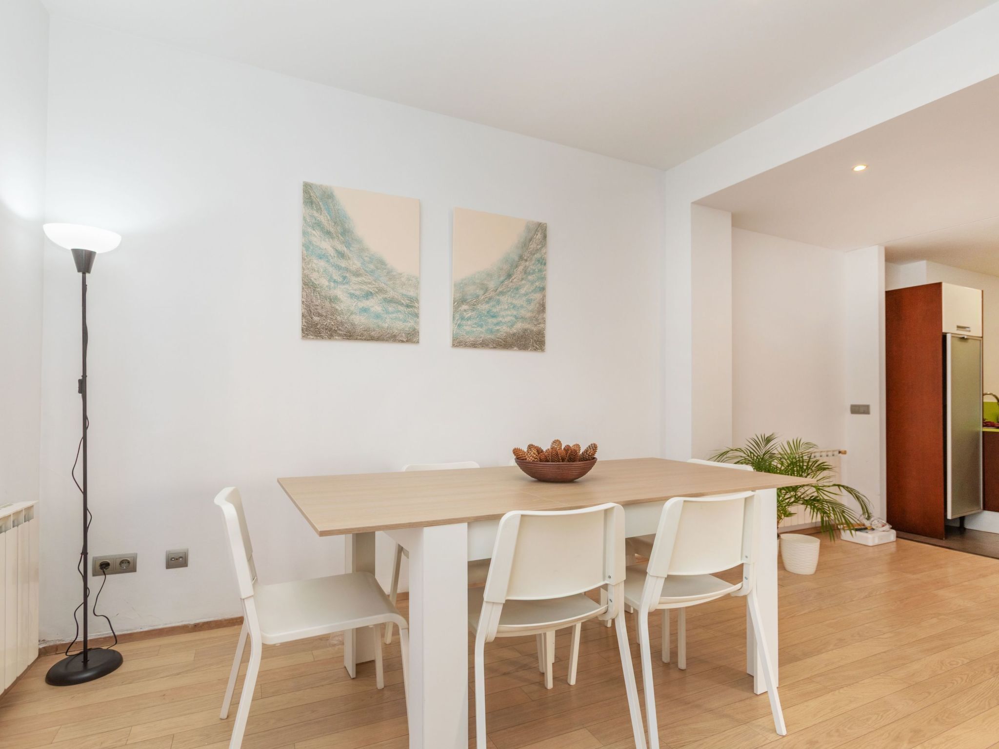 A dining area with a wooden table, white chairs, two abstract paintings on the wall, a floor lamp, and a potted plant.