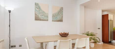 A dining area with a wooden table, white chairs, two abstract paintings on the wall, a floor lamp, and a potted plant.