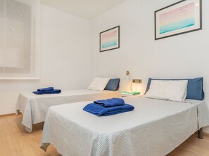 A hotel room with two beds, each with a striped bedspread and pillows, a bedside table with a lamp, and a window with a view. - Matilde by Interhome (St Antoni de Calonge)
