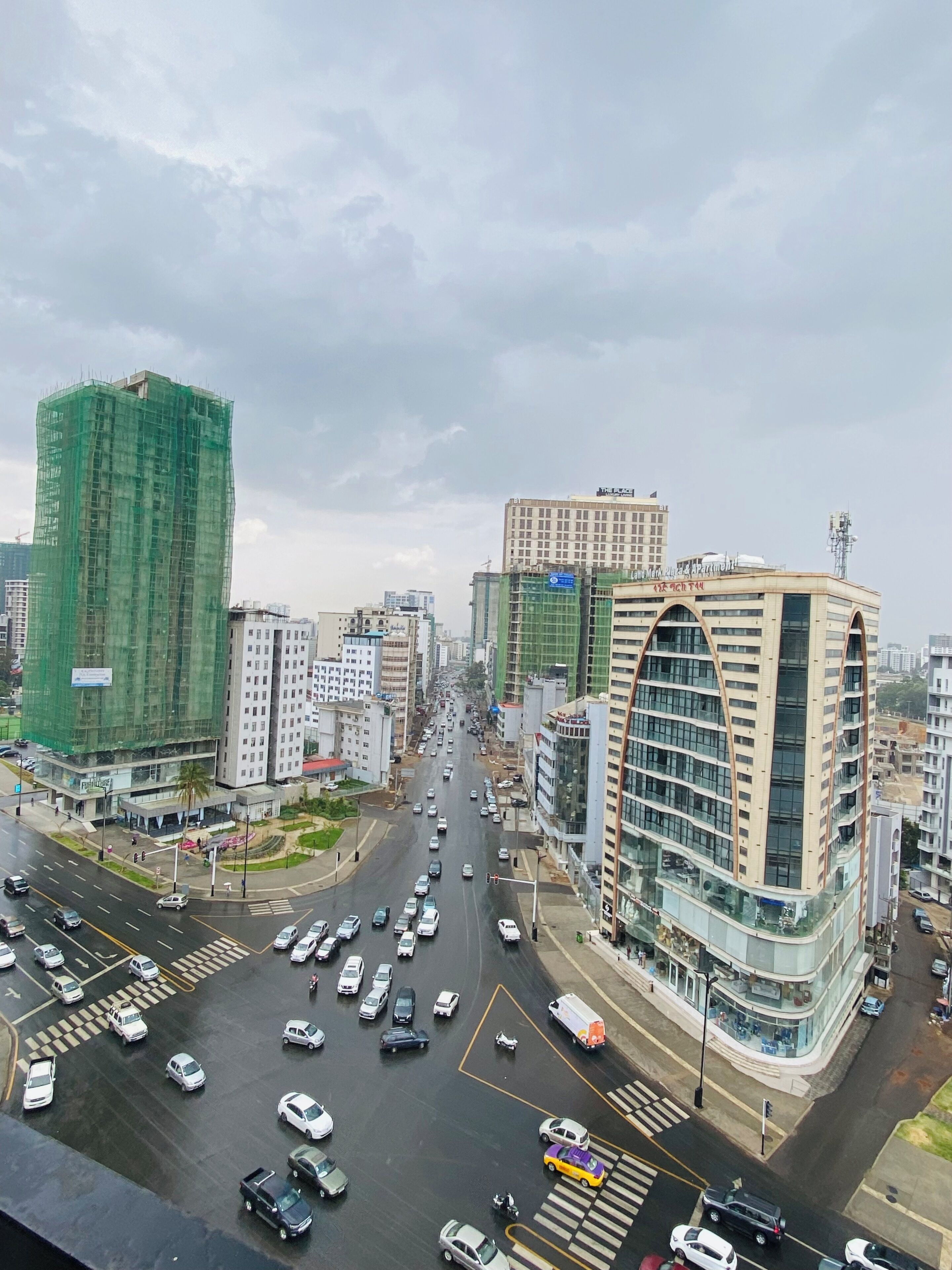 Un paisaje urbano con varios edificios, incluyendo un gran edificio verde en construcción, un edificio beige con fachada curva, y otros altos edificios.