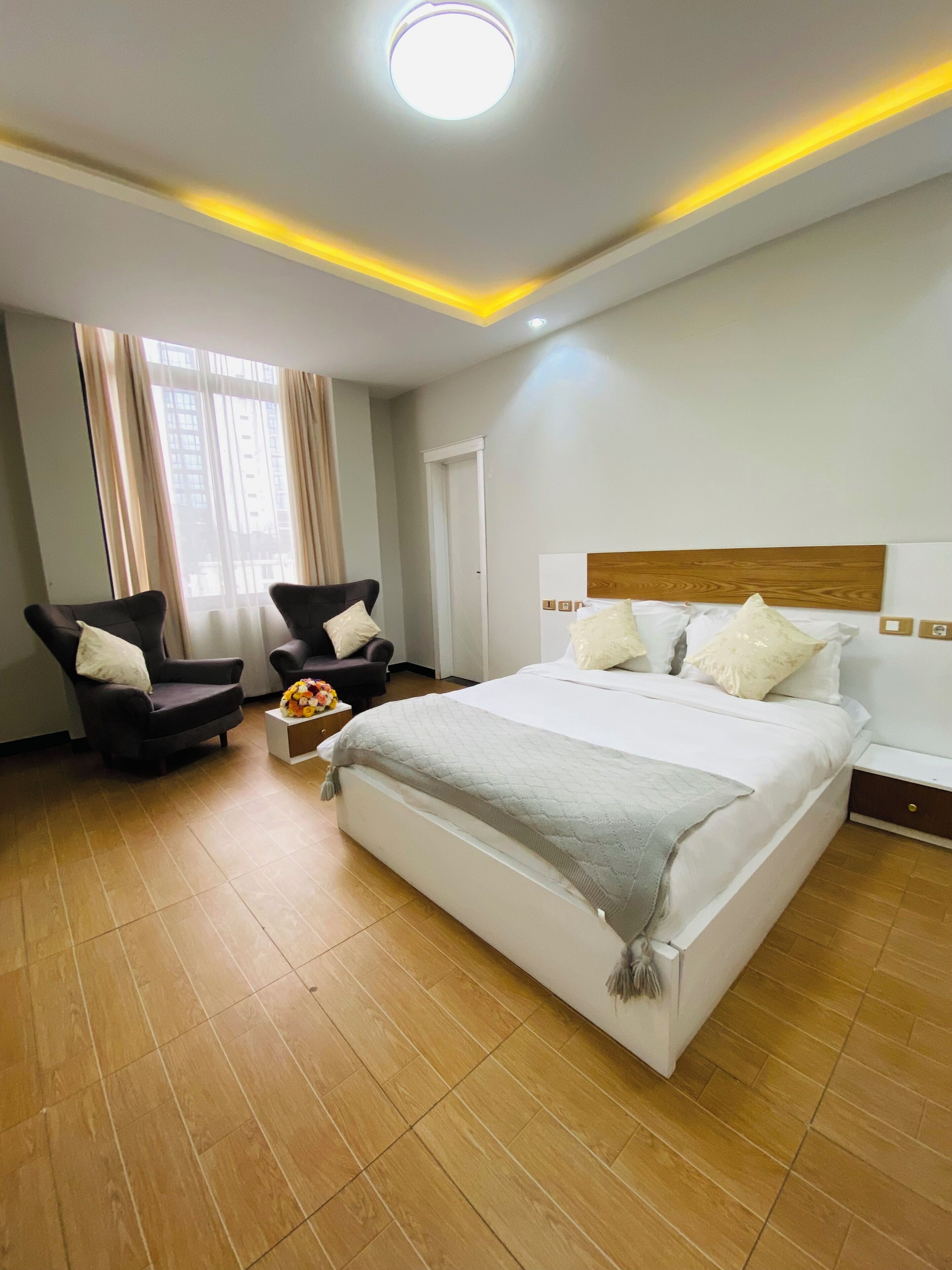 A hotel room with a large bed, two armchairs, a small table with a vase, and a view of buildings through the window.