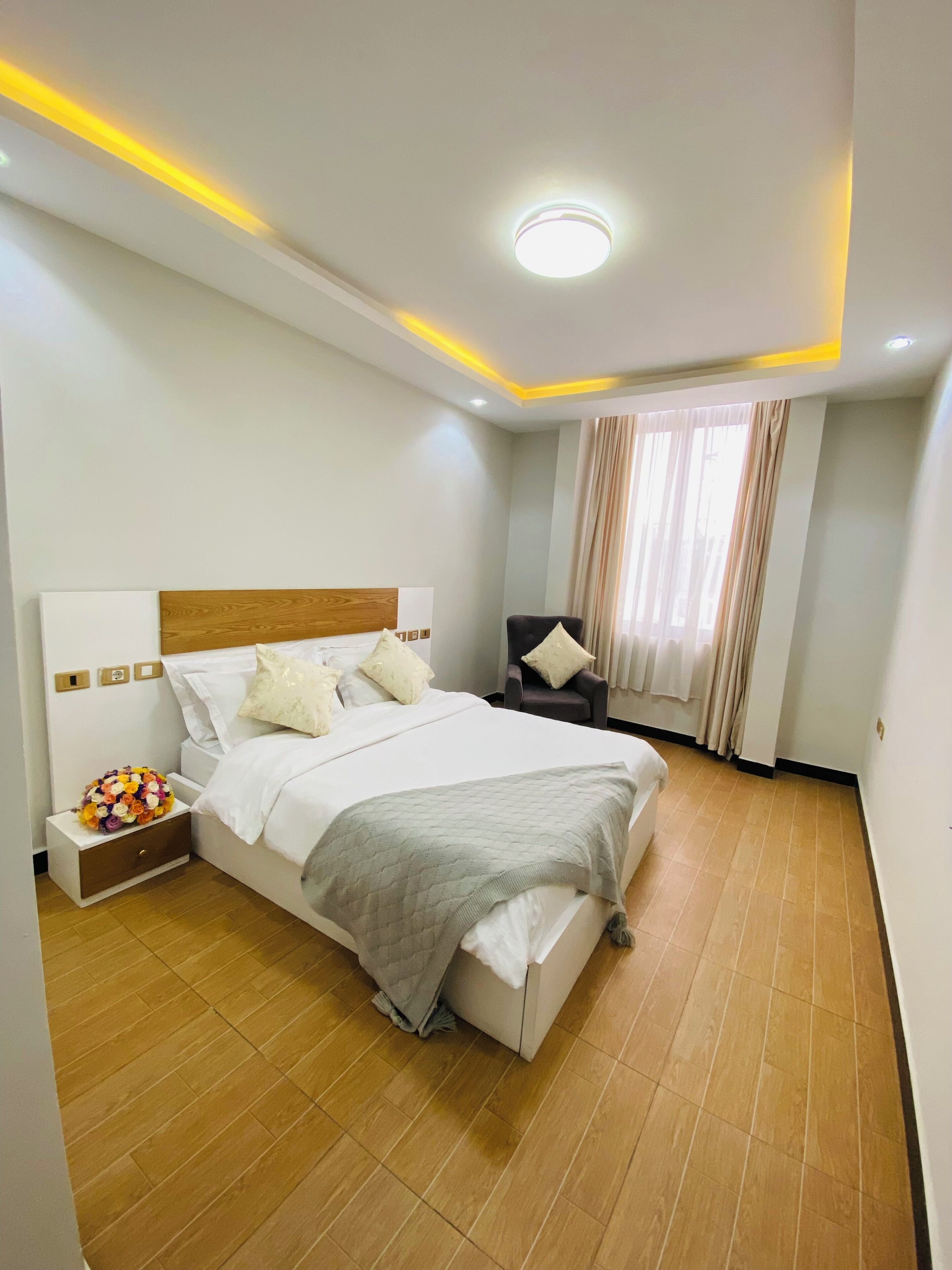 Moderni hotellihuone, jossa on suuri sänky, yöpöytä, jossa on kukka-asetelma, tuoli ja ikkuna, jossa on verhot.
