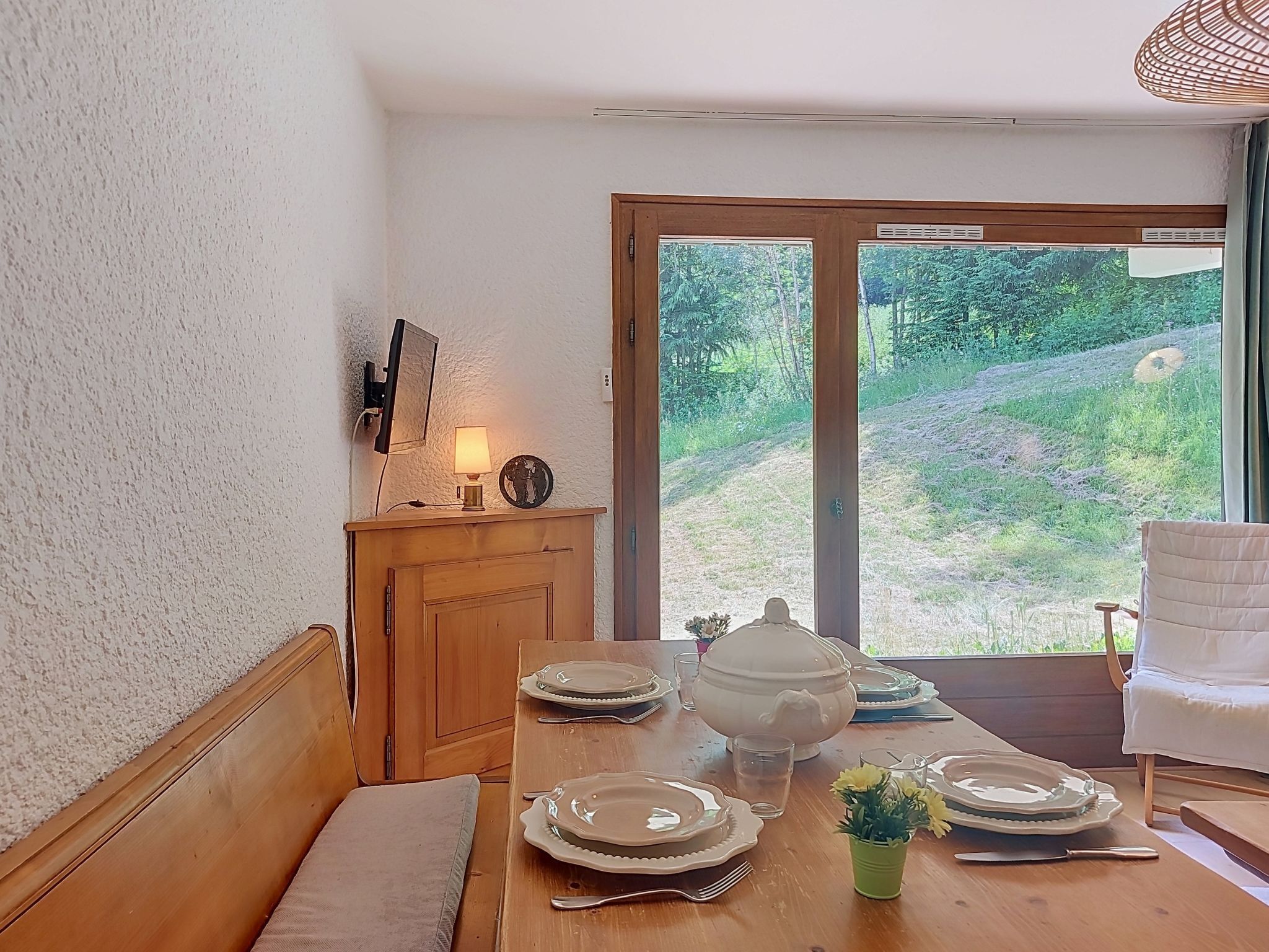 A dining area with a wooden table set for four, a wooden cabinet with a lamp, and a view of greenery through a glass door.