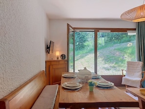 A dining area with a wooden table set for four, a bench, and a view of a green landscape through large windows. - Les Tétras by Interhome (Saint Gervais)