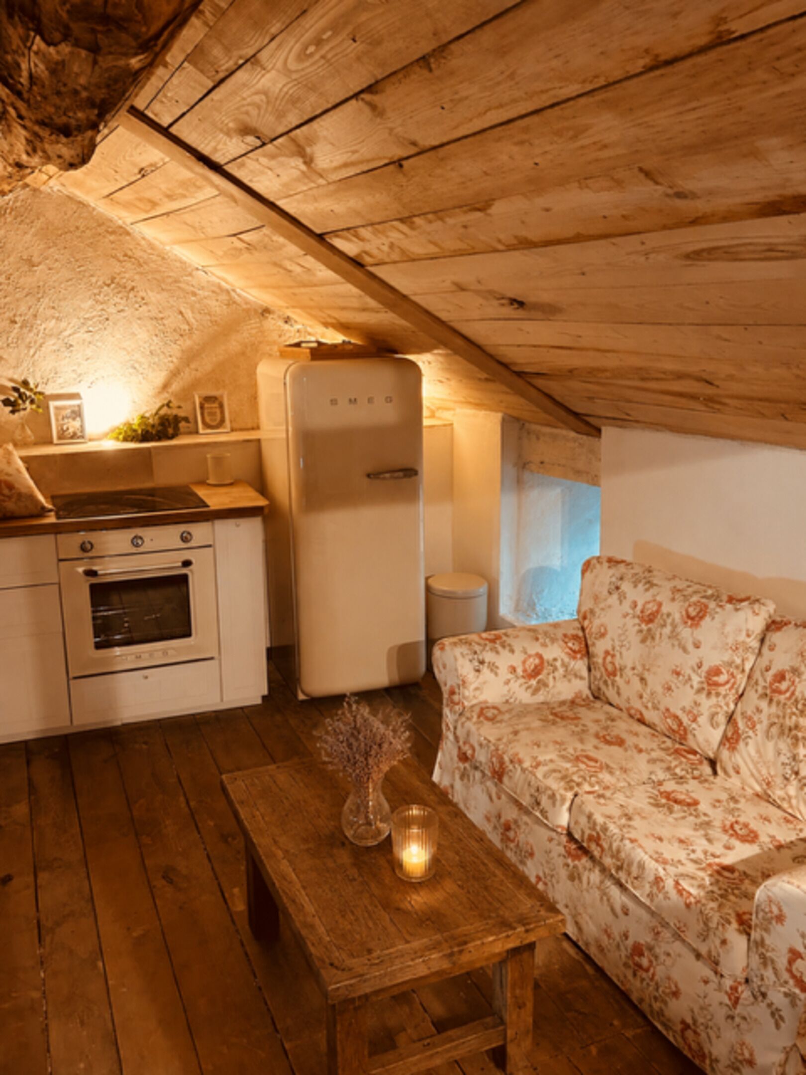Een gezellig slaapkamer onder het dak met houten balken, een kitchenette met fornuis en koelkast, een bloemenbank en een kleine houten tafel met een kaars.