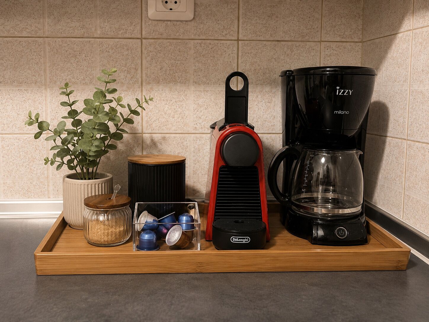 A kitchen counter with a coffee maker, a kettle, a plant, and a tray with jars and cups.