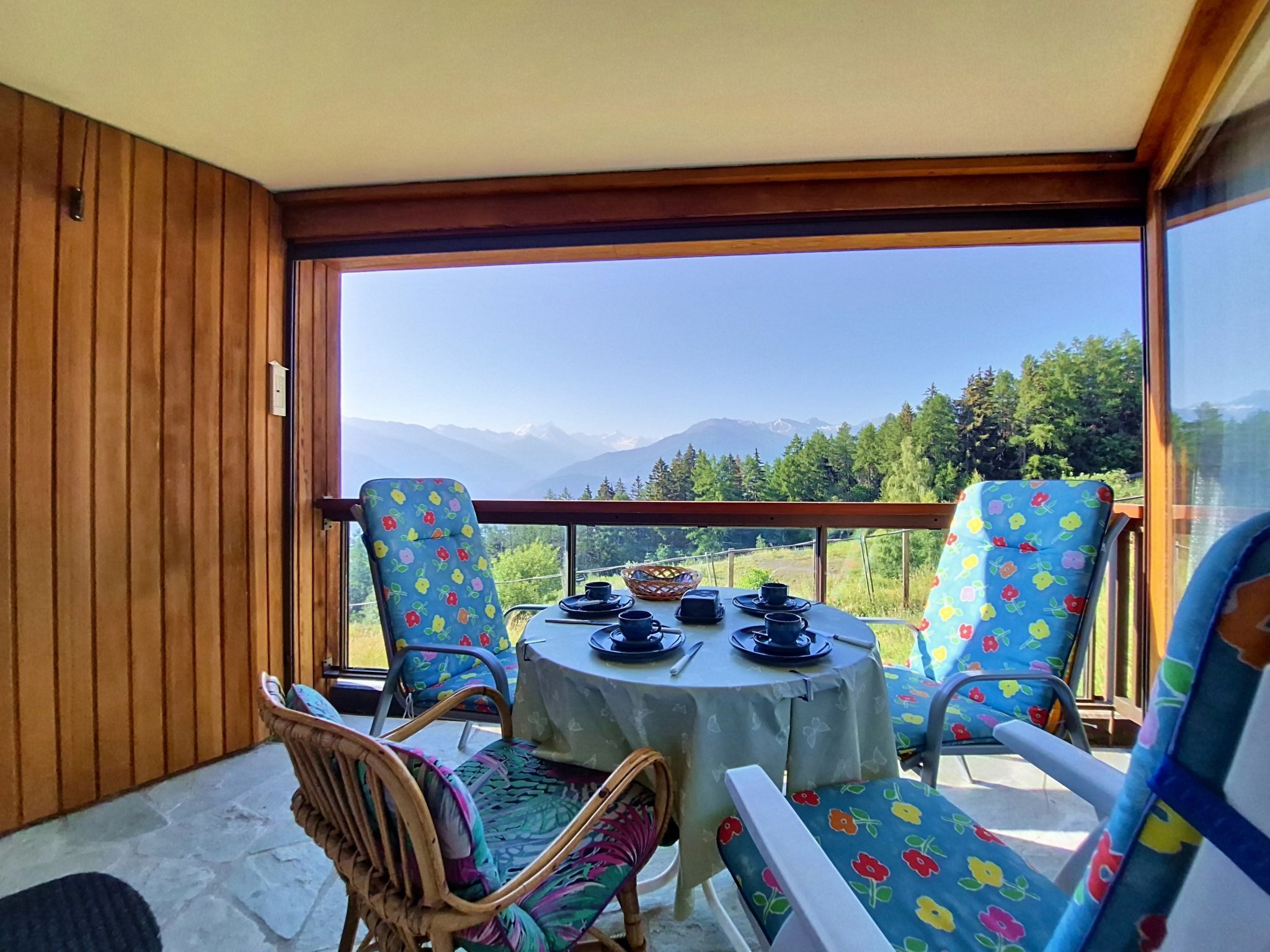 Une pièce avec une table dressée pour deux personnes, une chaise en osier et une vue sur les montagnes par de grandes fenêtres.