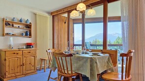 A dining area with a table set for four, wooden chairs, a wooden cabinet with shelves, and a view of mountains through large windows. - Mischabels 20 by Interhome (Crans-Montana)